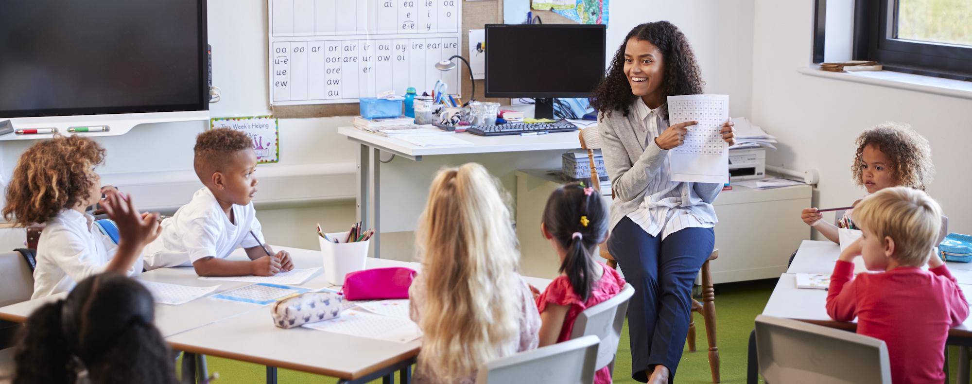 A teacheer sits in a chair in front of a classroom of kindergarteners. She is smiling and pointing to a chart in her hand.
