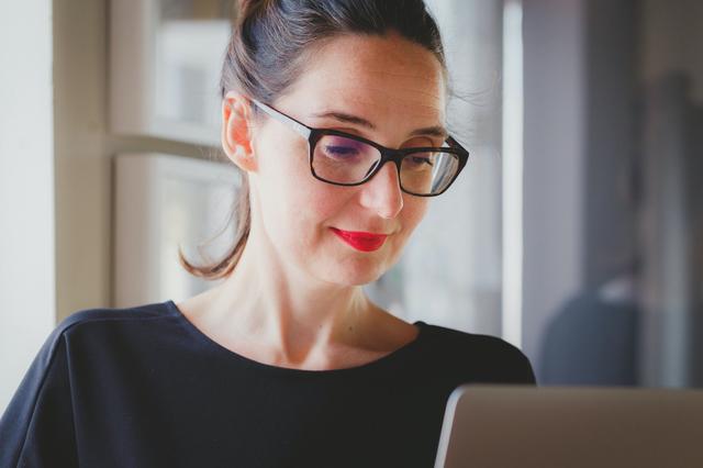 A woman with glasses works at a laptop and smiles slightly.