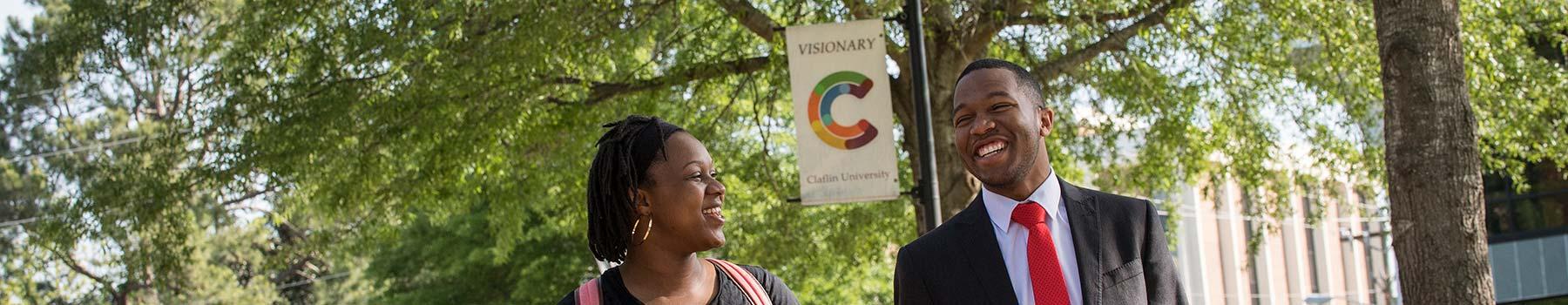 A man and a woman of color, smiling under the shade of trees in the Claflin University