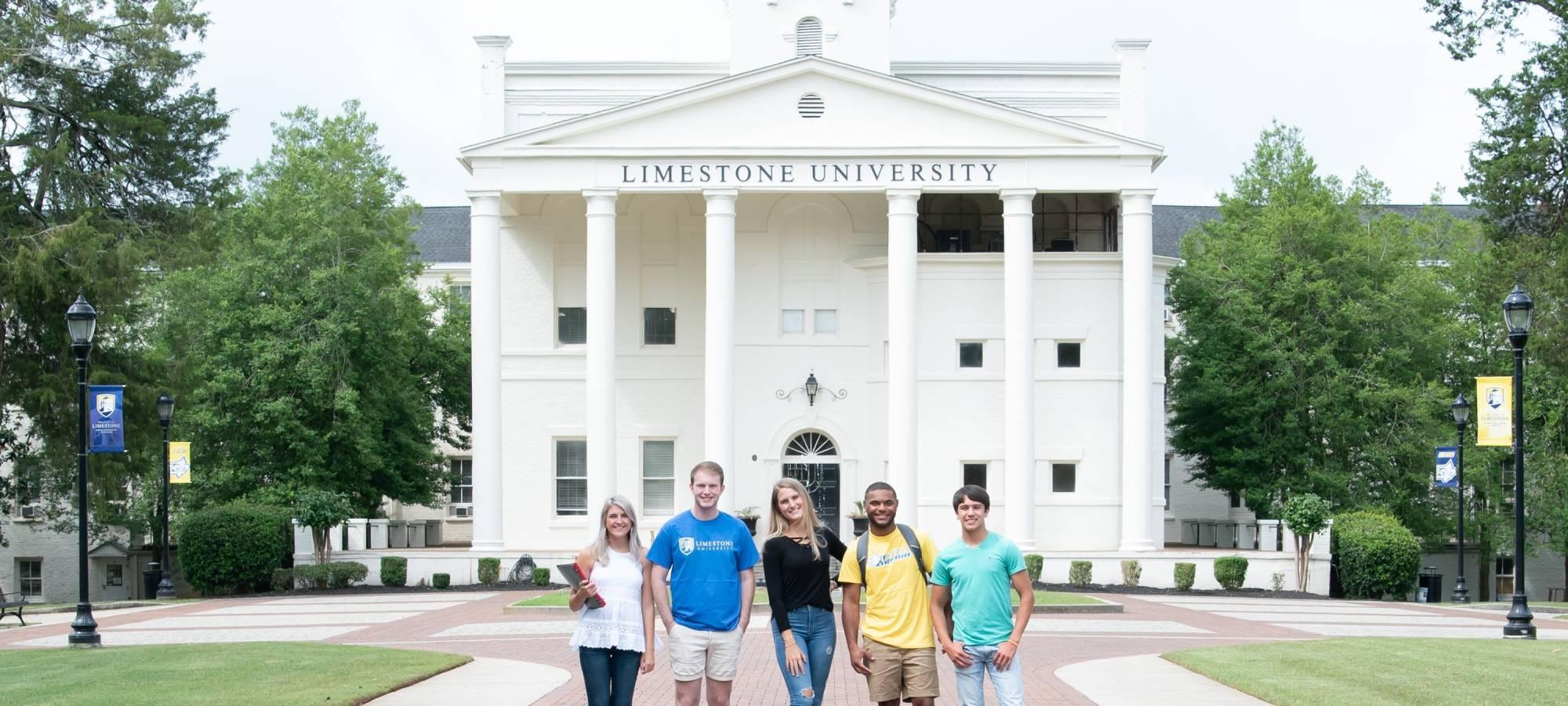Five smiling college students standing in front of school entrance.