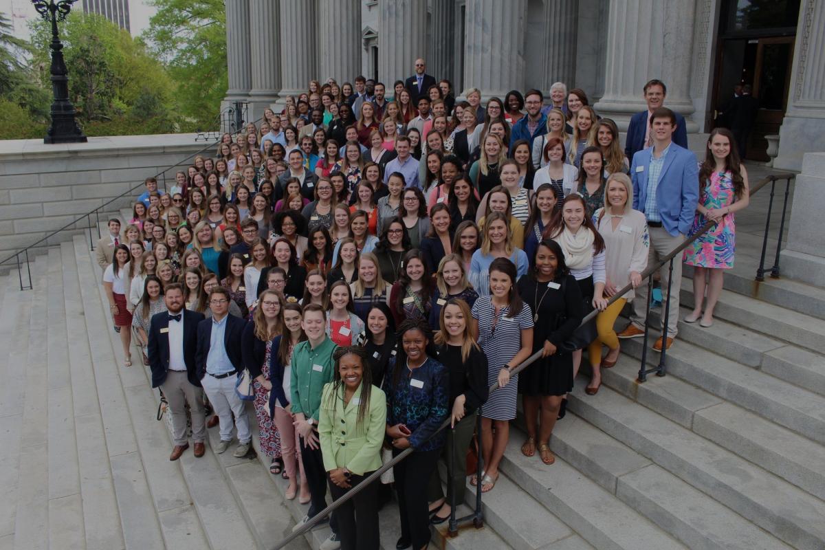 A large group of teaching fellows and mentors standing for a group photo on the stairs of a building with large white columns