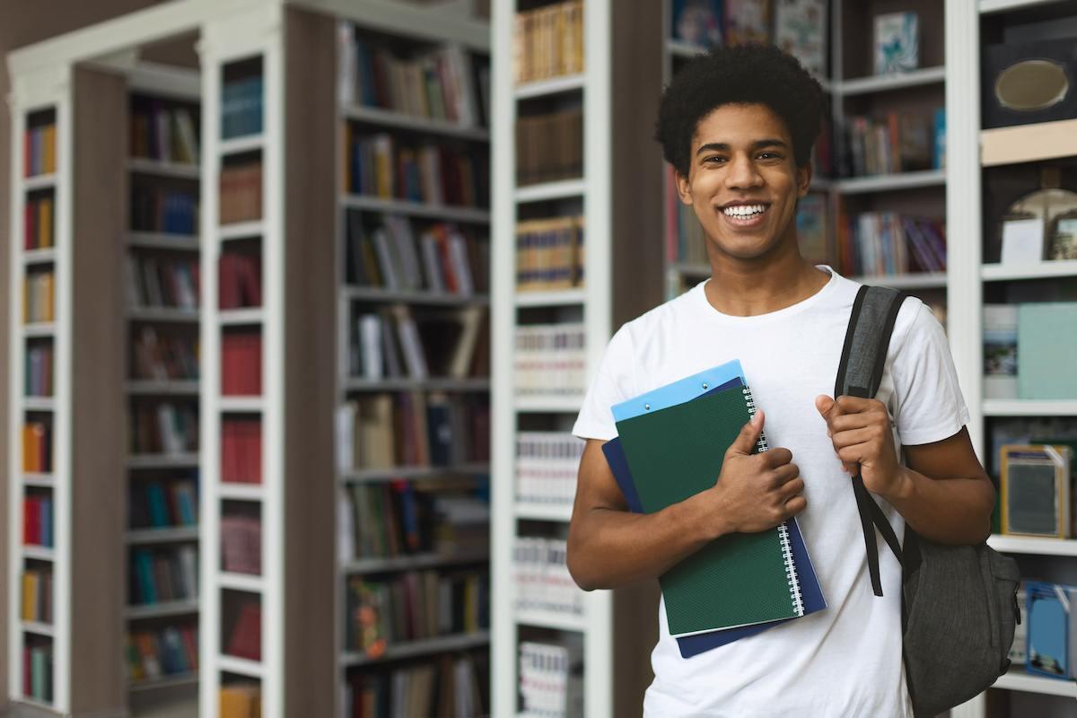 A future SC teacher standing in his campus library, holding books in one hand and a backpack on his shoulder with the other